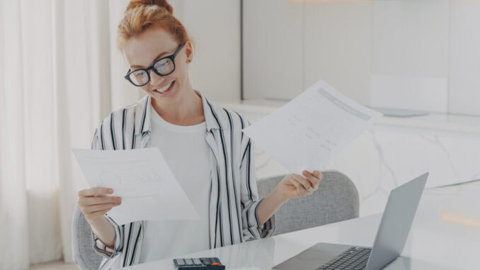Young excited redhead woman sitting at table at home, feeling happy after checking domestic bills,